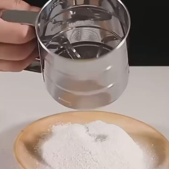 Demonstration video of stainless steel flour sifter with manual press handle, showing how to sift flour onto a plate for baking and cooking