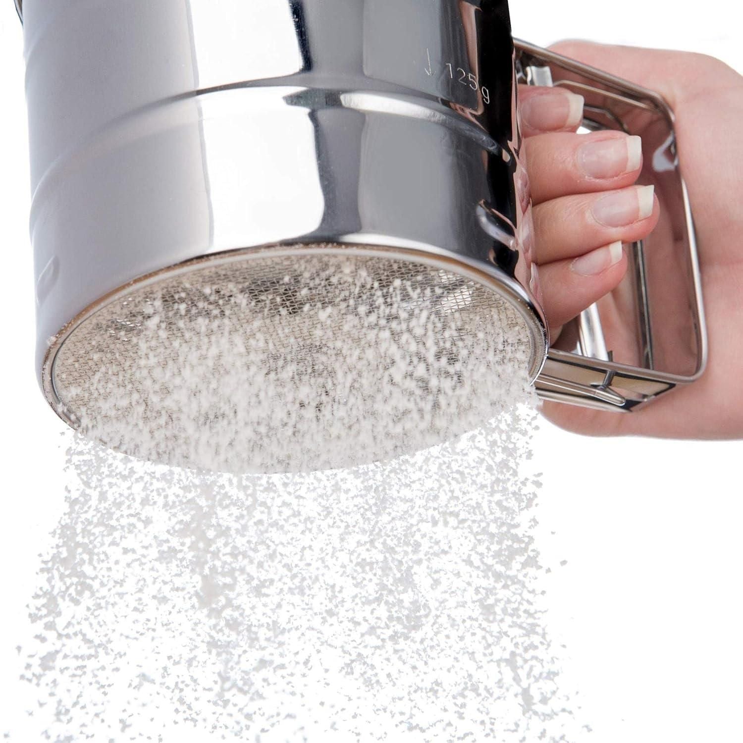 Close-up of stainless steel flour sifter with manual handle, sifting flour through a fine mesh for baking, perfect kitchen sieve for atta, sugar, cocoa, and powdered ingredients